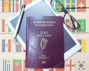 Irish passport placed on a surface with country flags in the background, alongside glasses and a pen.